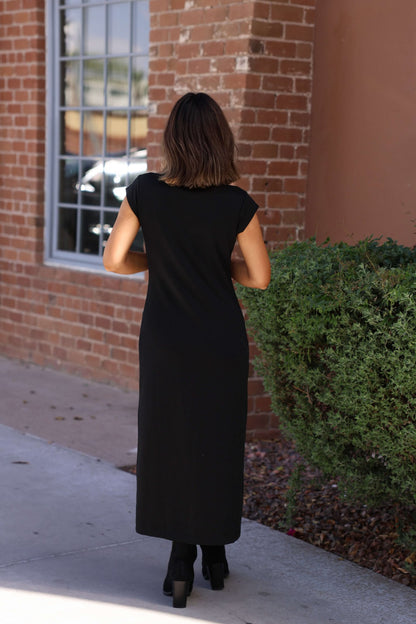 A woman in the Simple Timeless Black Midi Dress stands outside near a brick wall and green shrub, facing away.
