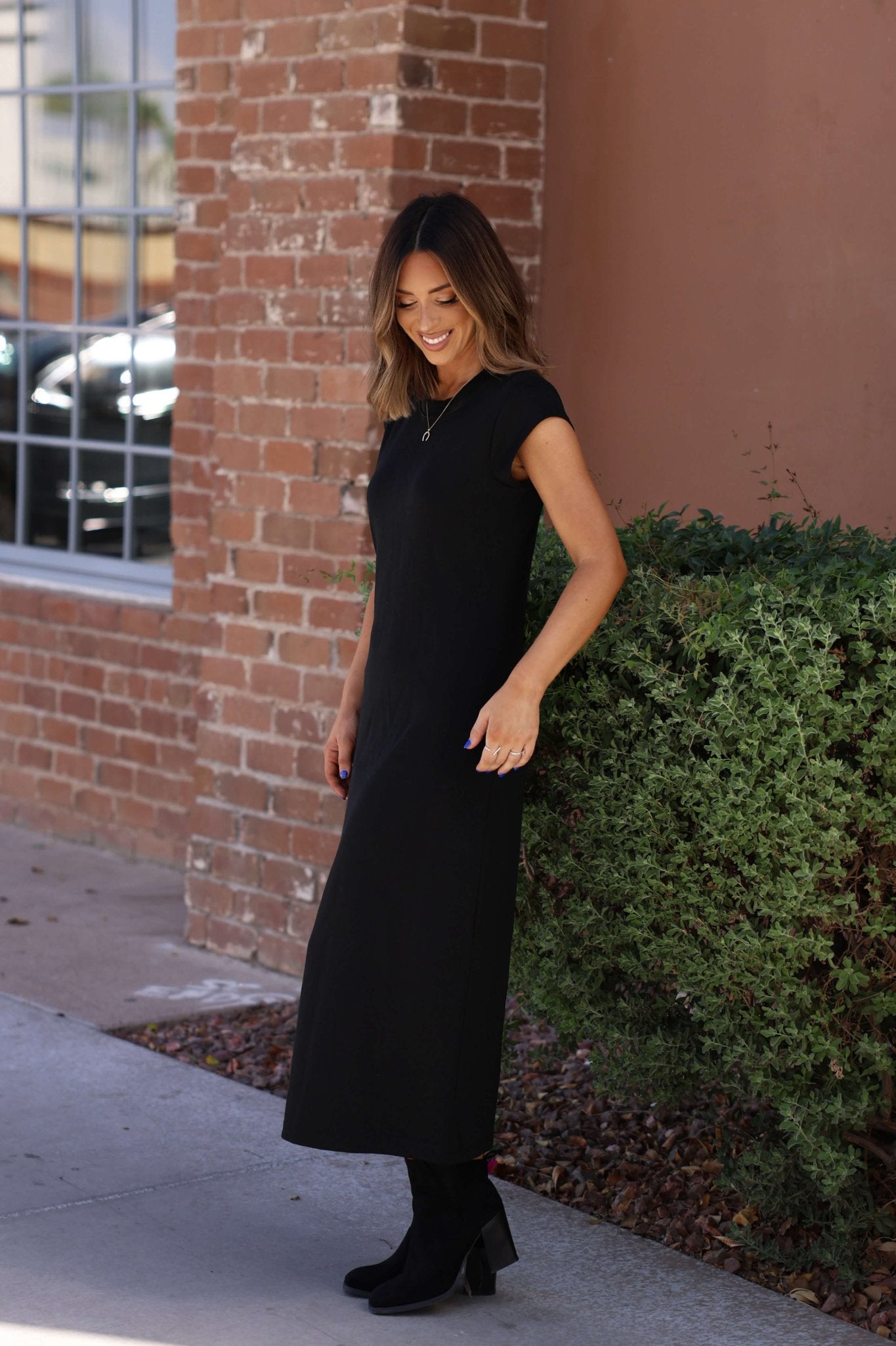 Smiling outside by a brick wall, she wears the Simple Timeless Black Midi Dress—a true wardrobe essential.