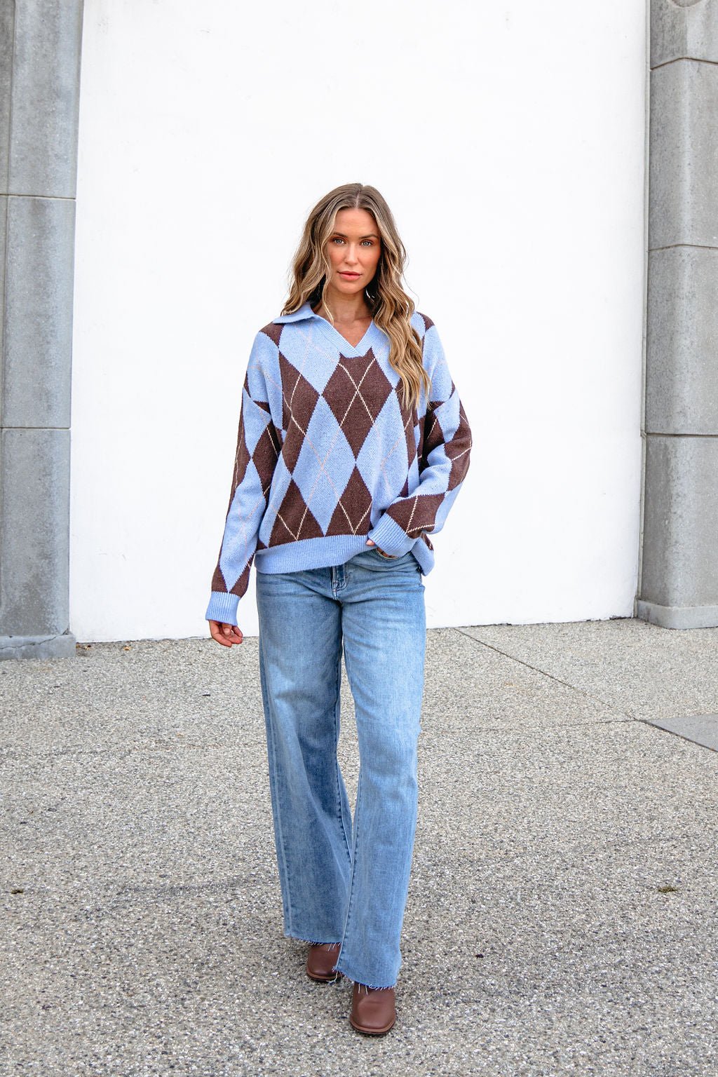 Woman in a Sky Blue Argyle Ribbed Sweater and light blue jeans stands by a white wall and stone columns.