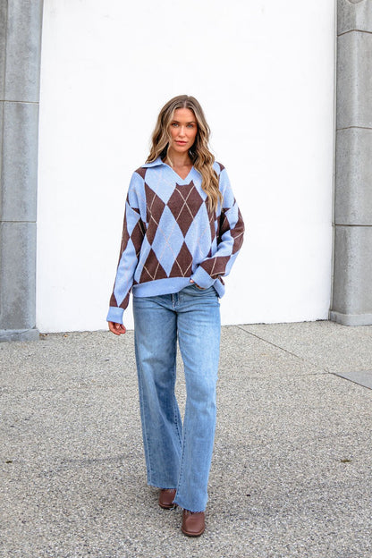 Woman in a Sky Blue Argyle Ribbed Sweater and light blue jeans stands by a white wall and stone columns.