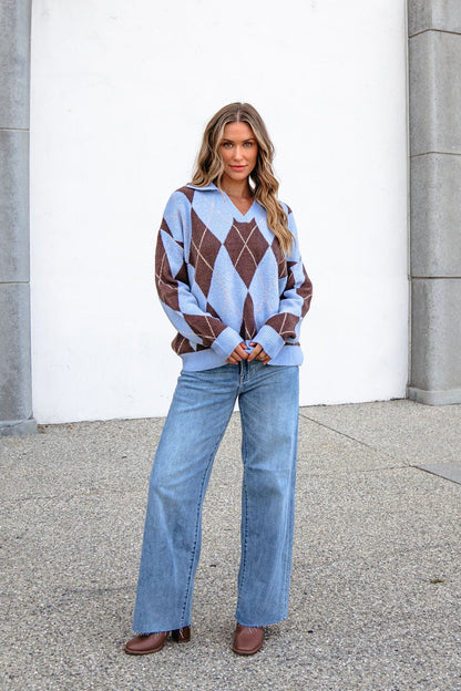 Woman in a Sky Blue Argyle Ribbed Sweater, wide-leg jeans, and brown shoes, standing outside by a white wall.