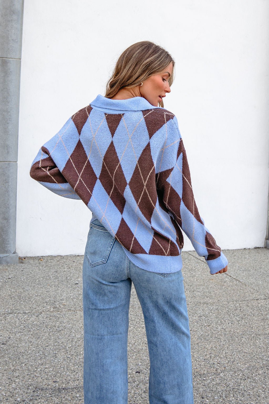 Woman facing away in a Sky Blue Argyle Ribbed Sweater with a collared V-neckline and light blue jeans on concrete.