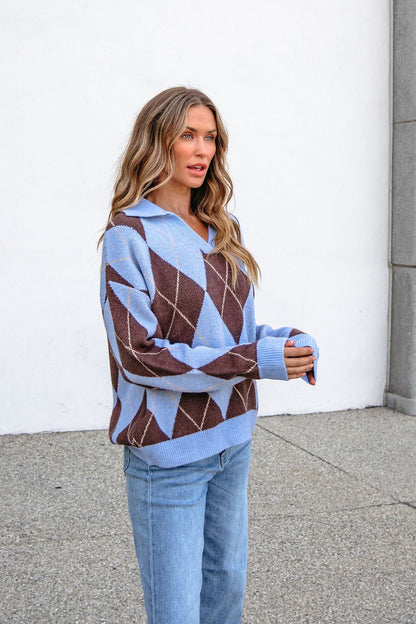 Woman wearing a Sky Blue Argyle Ribbed Sweater with jeans stands outdoors by a white wall, gazing to the side.