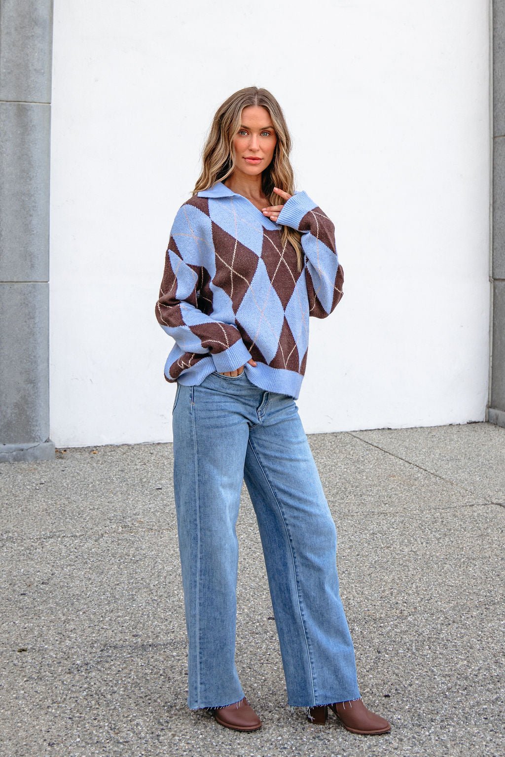Woman in a Sky Blue Argyle Ribbed Sweater, blue jeans, and brown boots stands outdoors against a white wall.