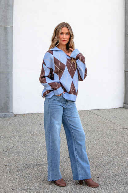 Woman in a Sky Blue Argyle Ribbed Sweater, blue jeans, and brown boots stands outdoors against a white wall.