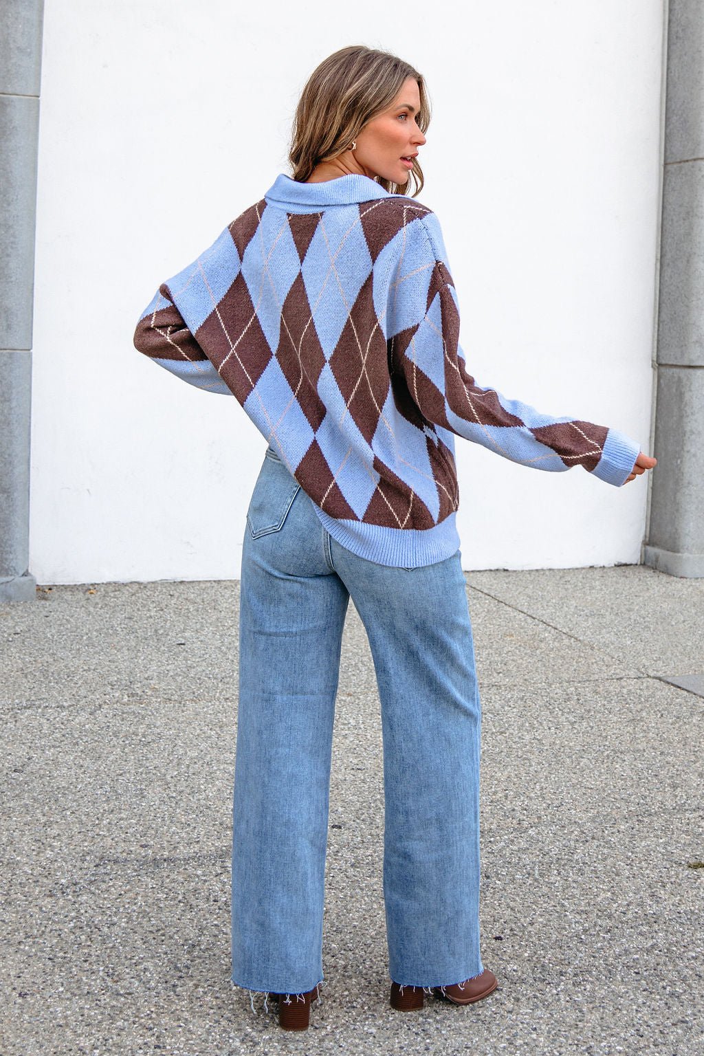 Someone in the Sky Blue Argyle Ribbed Sweater and light blue jeans stands outdoors facing away from the camera.
