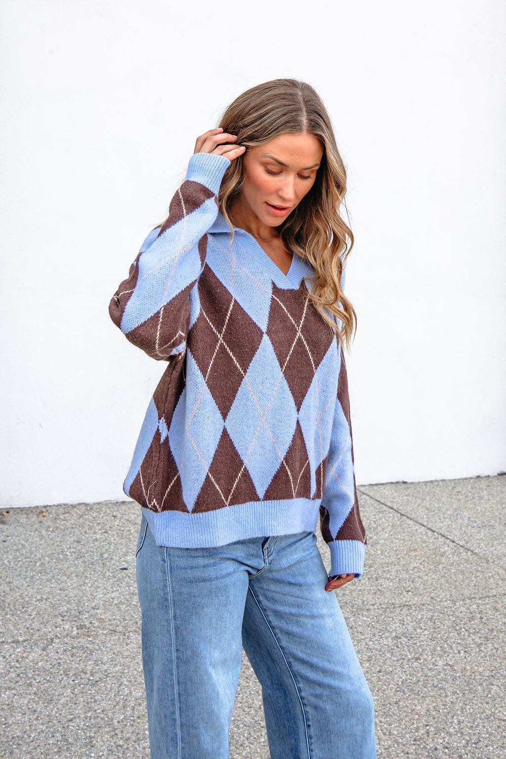 Woman in a Sky Blue Argyle Ribbed Sweater with a collared V-neck stands outdoors on concrete, looking down and touching her hair.