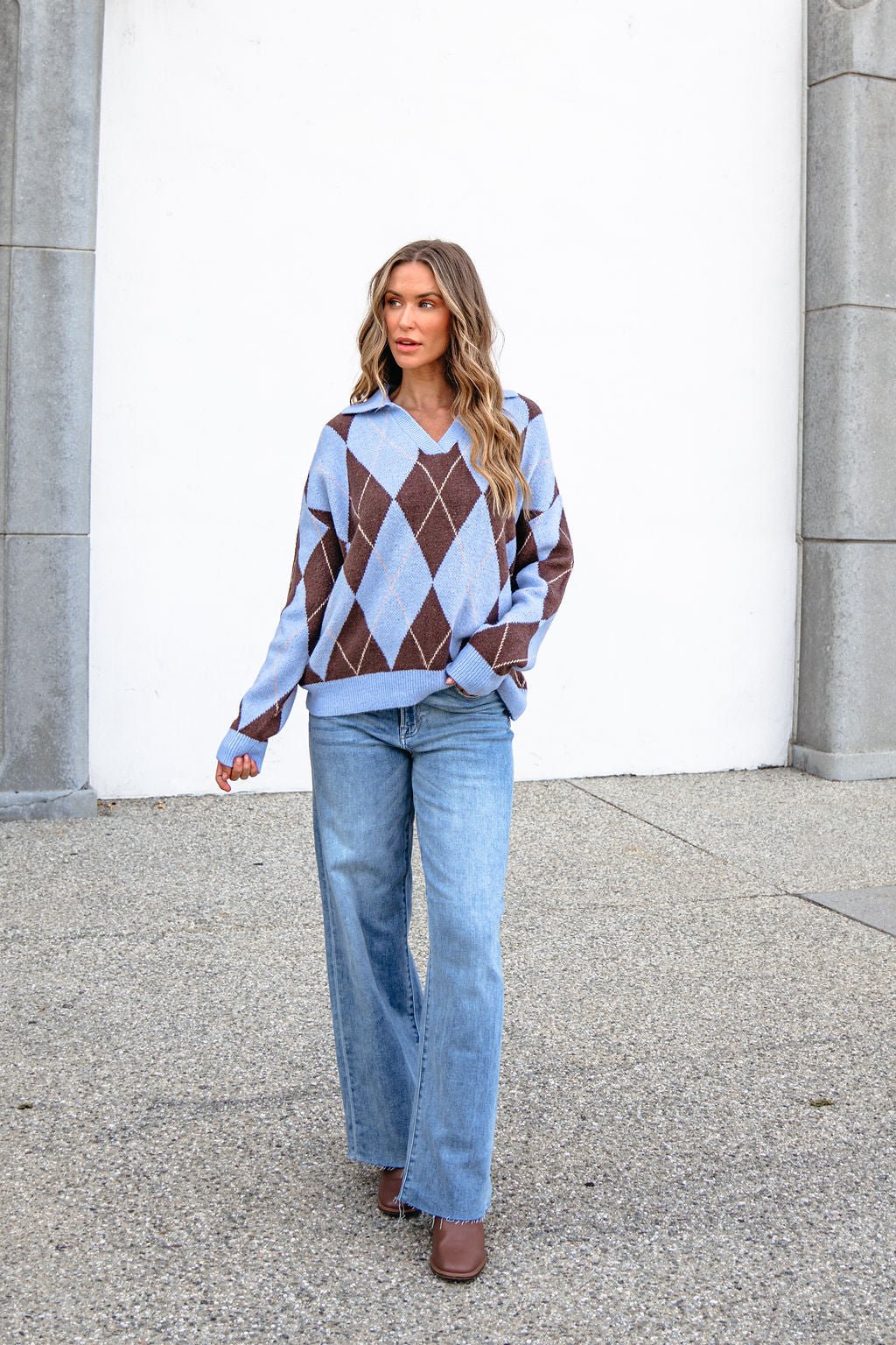 Woman with wavy hair in a Sky Blue Argyle Ribbed Sweater and light blue jeans walks outdoors by a white wall.