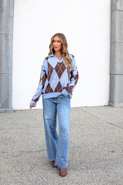 Woman with wavy hair in a Sky Blue Argyle Ribbed Sweater and light blue jeans walks outdoors by a white wall.