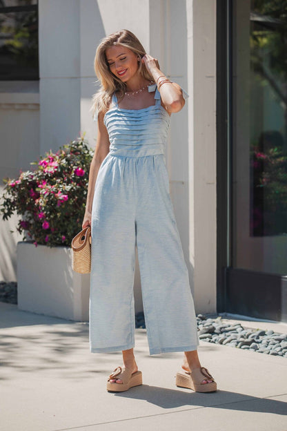 Woman in a Skylar Pleated Wide Leg Jumpsuit - Light Blue stands outdoors holding a clutch, with flowers and sunlight behind her.