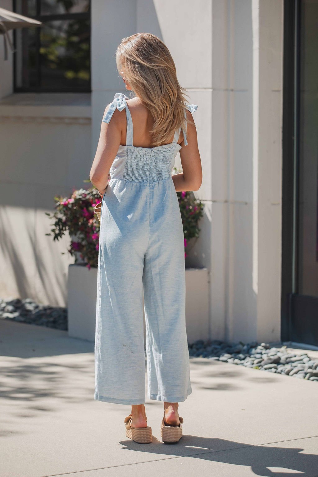 Woman wears a Skylar Pleated Wide Leg Jumpsuit in light blue and platform sandals, standing outdoors near flowers with her back to the camera.