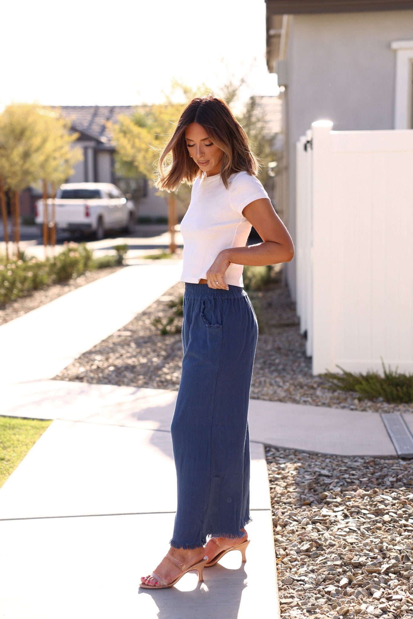 Woman in a white t-shirt and Slate Blue Frayed Linen Wide Leg Pants stands outdoors, looking down and smiling.
