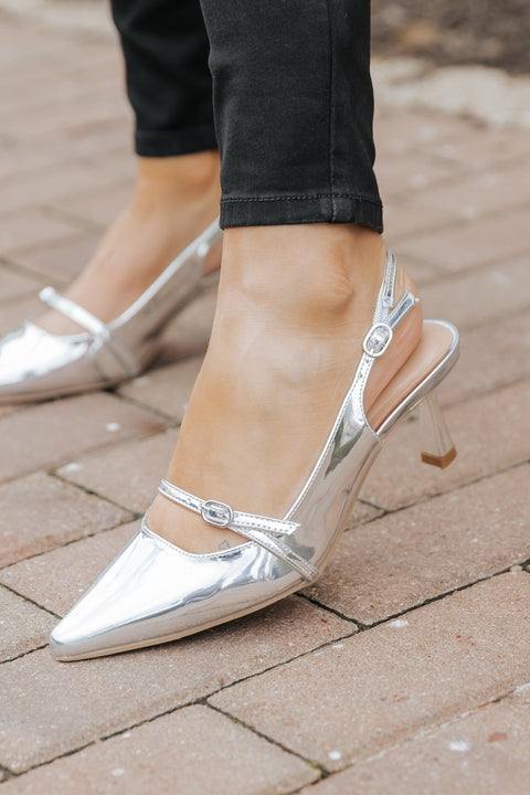 Someone in black pants and Sleek Silver Buckle Detail Slingback Heels stands on a brick pavement.