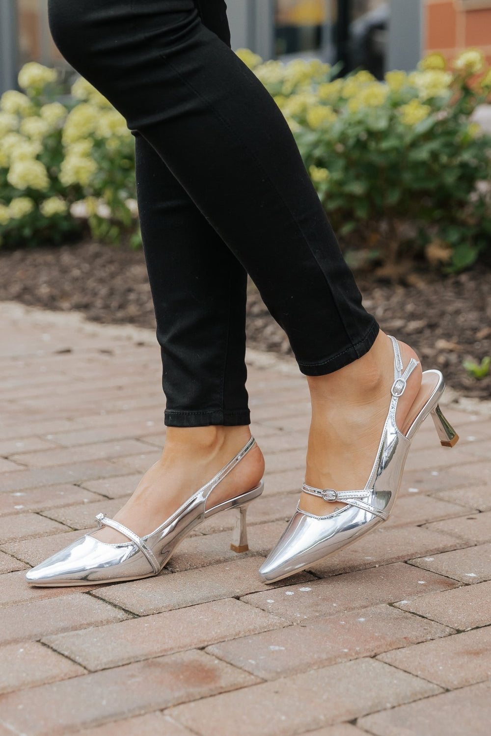 Person in Sleek Silver Buckle Detail Slingback Heels - FINAL SALE and black pants stands on a brick path with greenery behind.