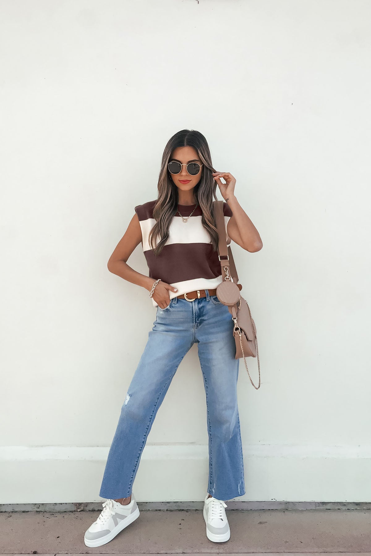 Woman in a Sleeveless Brown and Cream Striped Sweater - DOORBUSTER, blue jeans, and accessories poses against a white wall.