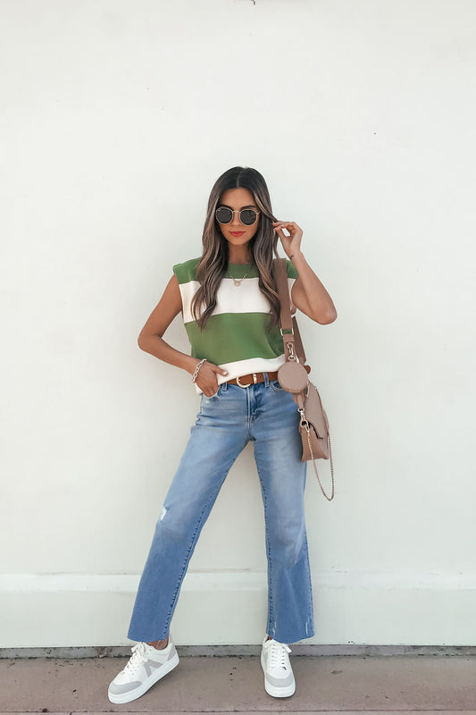 Woman in sunglasses wears a Sleeveless Green and Cream Striped Sweater, blue jeans, white sneakers, and a beige crossbody bag.
