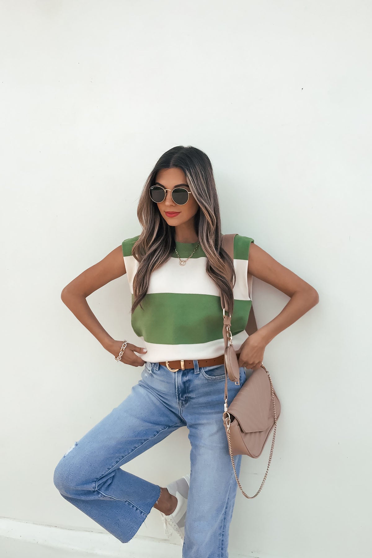 Woman in sunglasses poses against a white wall, wearing a Sleeveless Green and Cream Striped Sweater, blue jeans, and a beige purse.