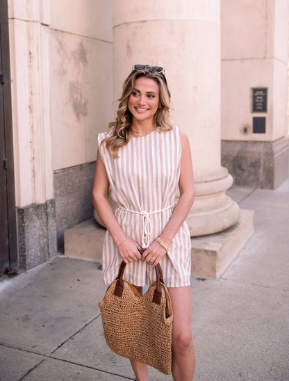 Woman wearing the Sleeveless Mocha Striped Belted Romper and holding a woven bag, smiling on a city sidewalk by columns.