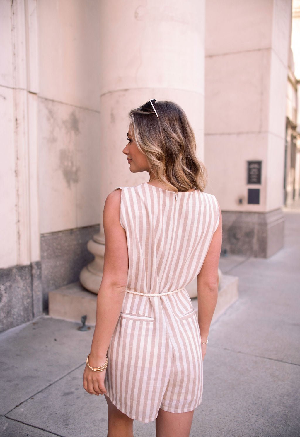 A woman with wavy blonde hair wears a Sleeveless Mocha Striped Belted Romper, sunglasses, and stands outdoors facing away.