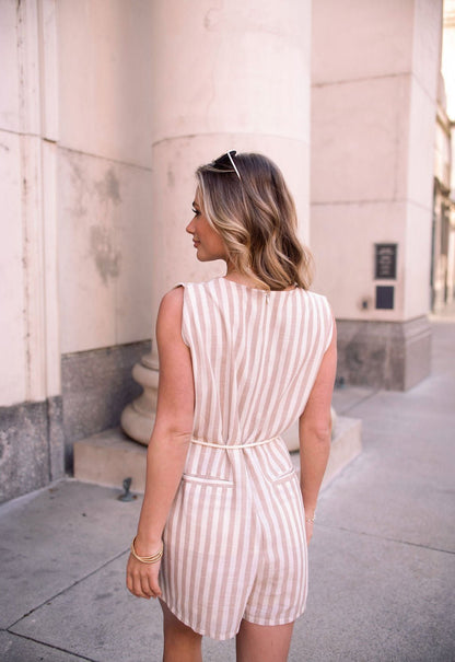 A woman with wavy blonde hair wears a Sleeveless Mocha Striped Belted Romper, sunglasses, and stands outdoors facing away.