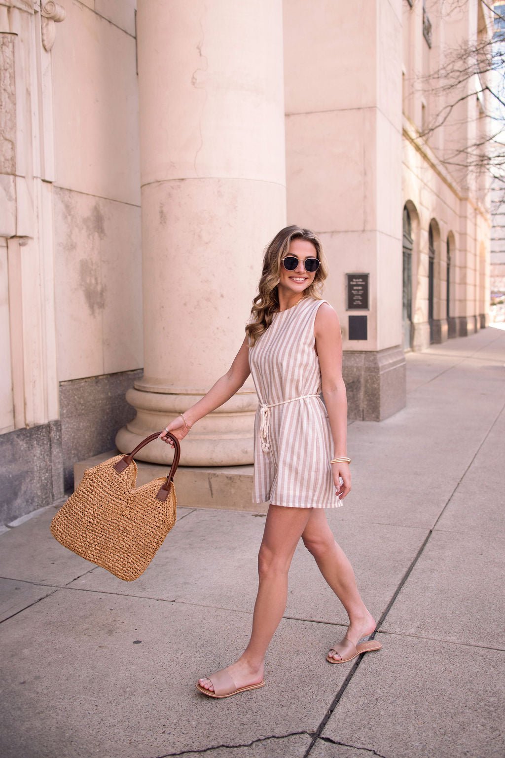 Woman wearing a Sleeveless Mocha Striped Belted Romper, holding a woven tote bag, walking on a city sidewalk.