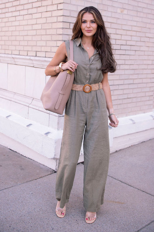 Woman in a Sleeveless Olive Belted Linen Jumpsuit with a tan belt and beige bag stands on a city sidewalk by a brick wall.