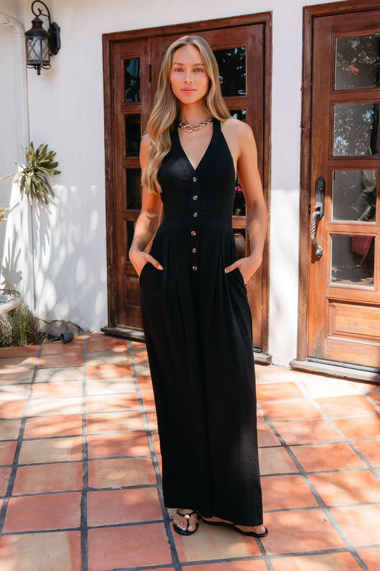 Woman in Sloane Halter Wide Leg Jumpsuit - Black, standing on a tiled patio before wooden doors, wearing sandals.
