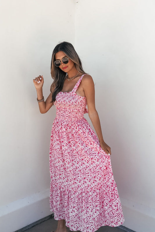 Woman in sunglasses wears the Smocked Bow Tie Print Midi Dress - Pink, standing against a white wall and holding the skirt.