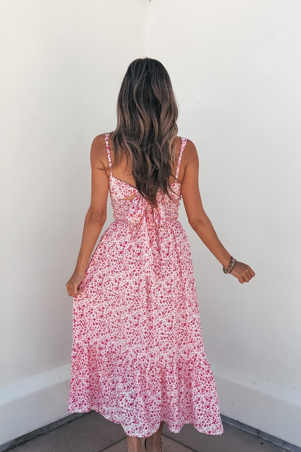 A woman with long wavy hair models the Smocked Bow Tie Print Midi Dress - Pink, facing away from the camera against a white wall.