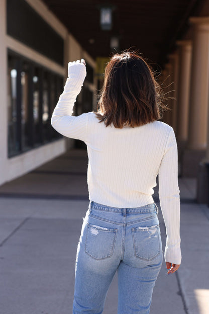 A woman in blue jeans and the Smoky Dawn Ribbed Sweater in Cream walks outside with her back to the camera, raising her hand.