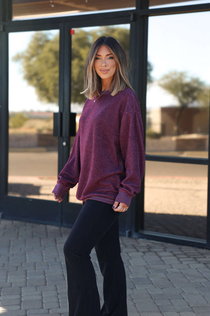 A woman with shoulder-length hair smiles outside glass doors, wearing a Soft Brushed Oversized Sweatshirt in plum with black pants.