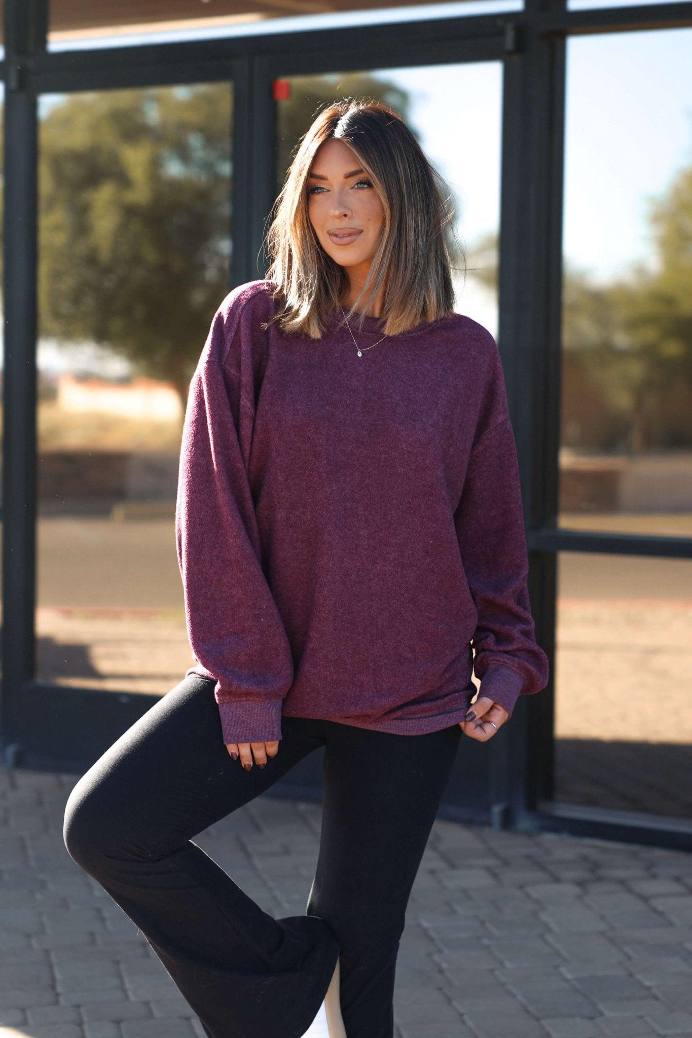 A woman with shoulder-length hair wears the Soft Brushed Oversized Sweatshirt in plum and black pants outdoors on a sunny day.