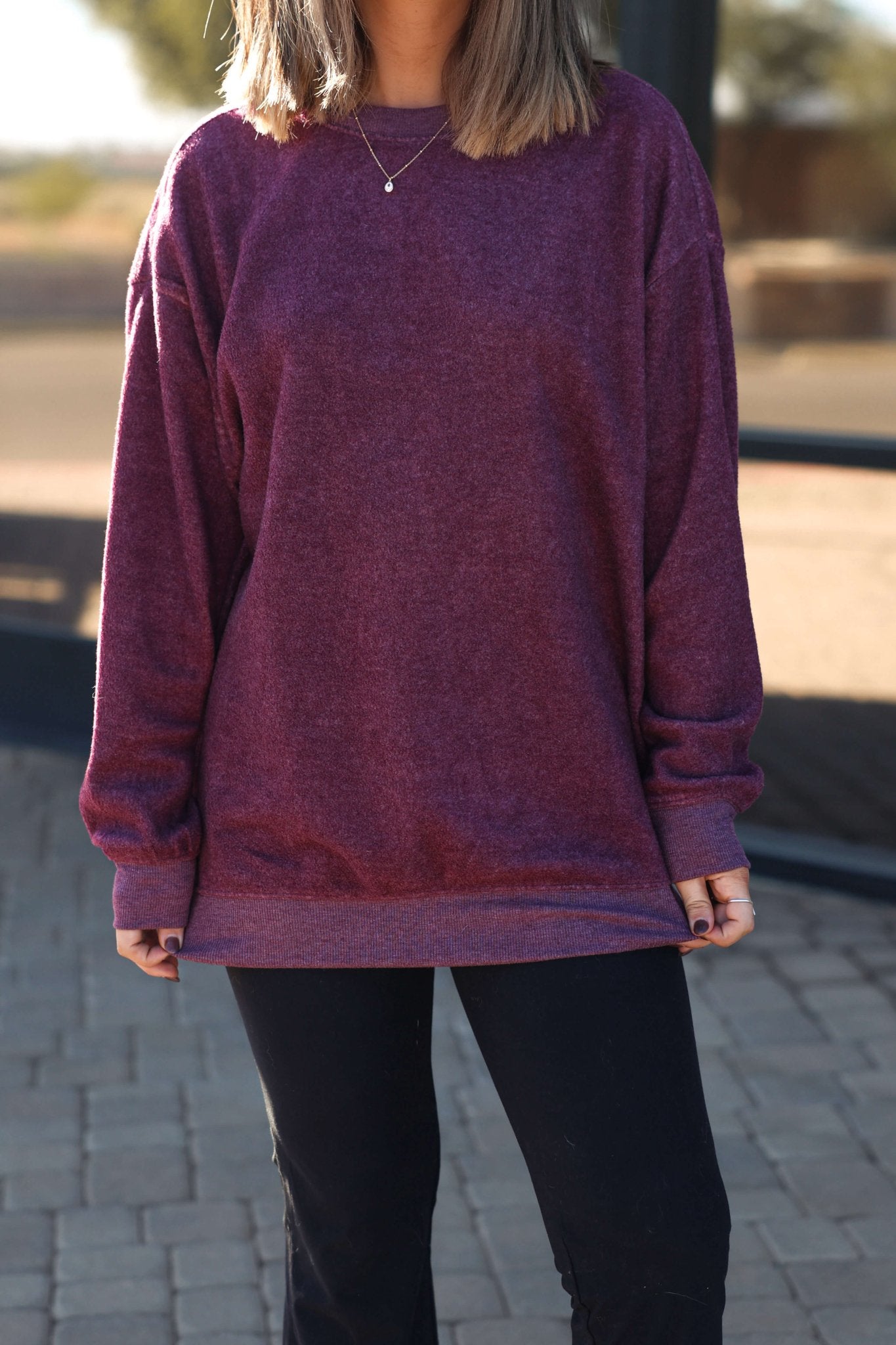 A woman stands outdoors on a sunny day wearing a Soft Brushed Oversized Sweatshirt in plum, black pants, and a necklace.