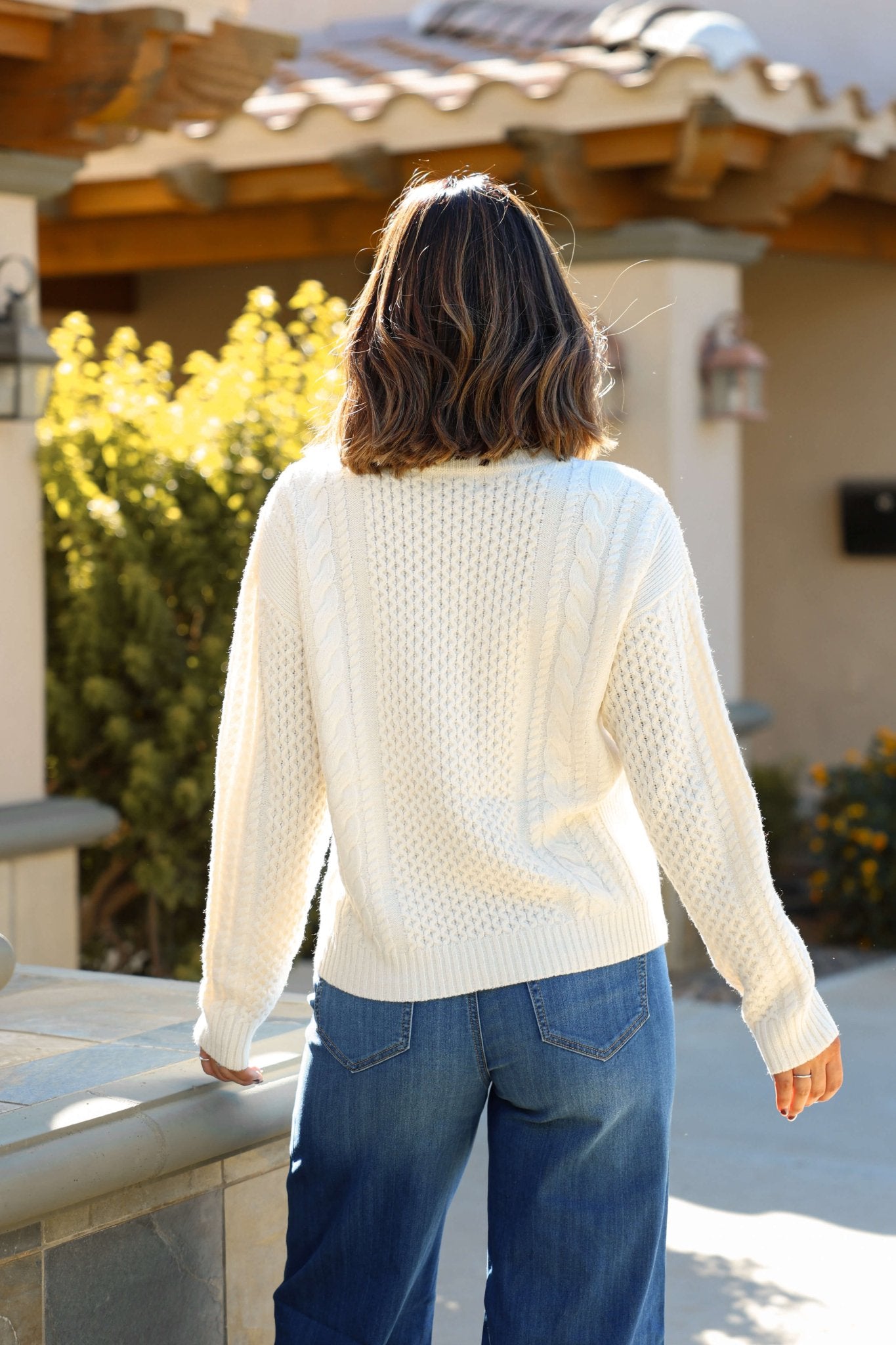 A person in a Soft Cream Cable Knit Sweater and blue jeans stands outdoors with shoulder-length hair, facing away from the camera.