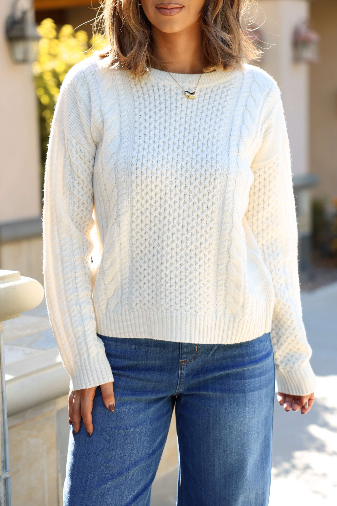 Woman in a Soft Cream Cable Knit Sweater and blue jeans stands outdoors, sunlight illuminating her hair and the scene.