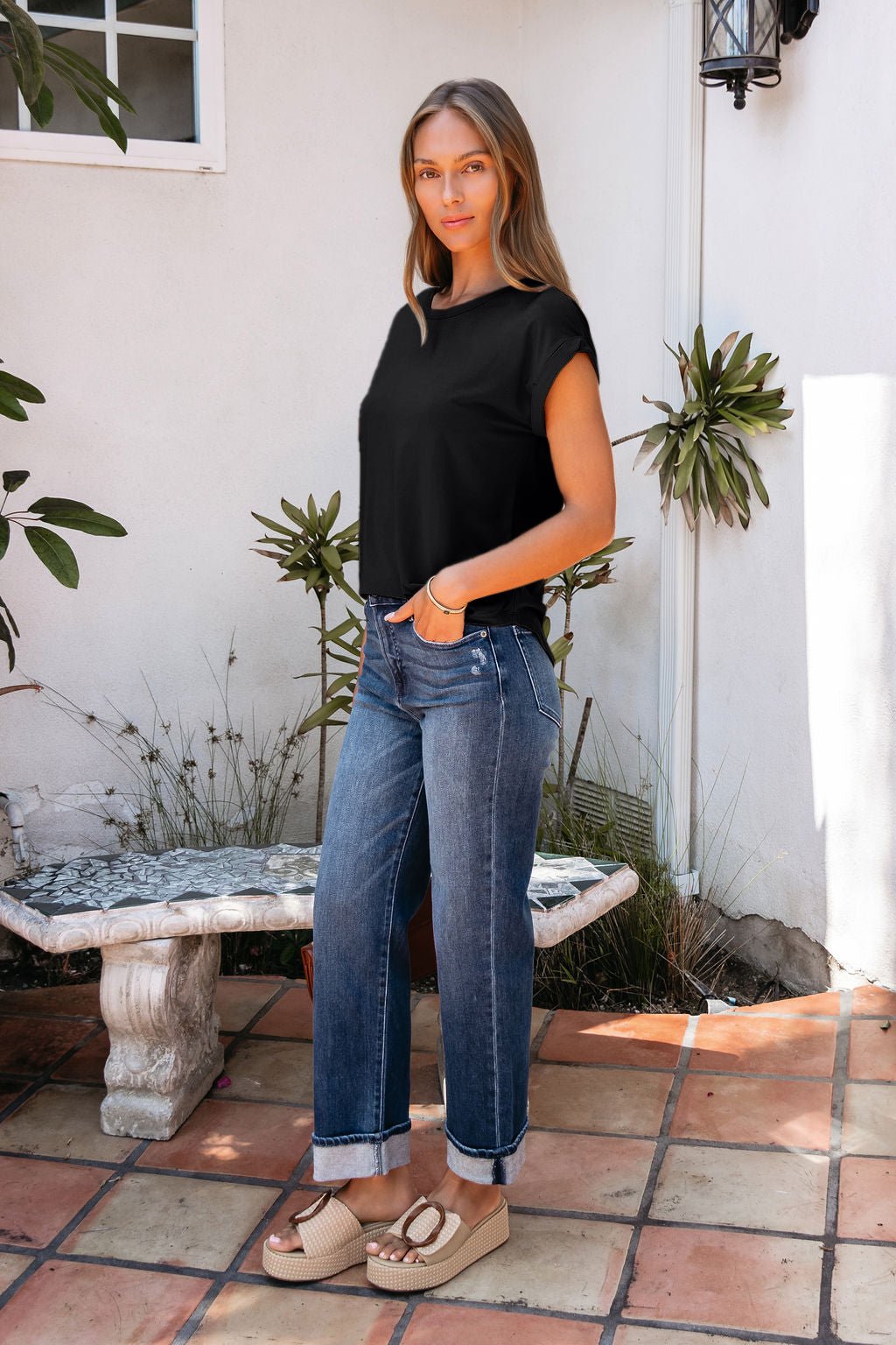 Woman standing on a tiled patio in a relaxed fit Soft Modal Basic Tee - Black, cuffed jeans, and platform sandals near plants.