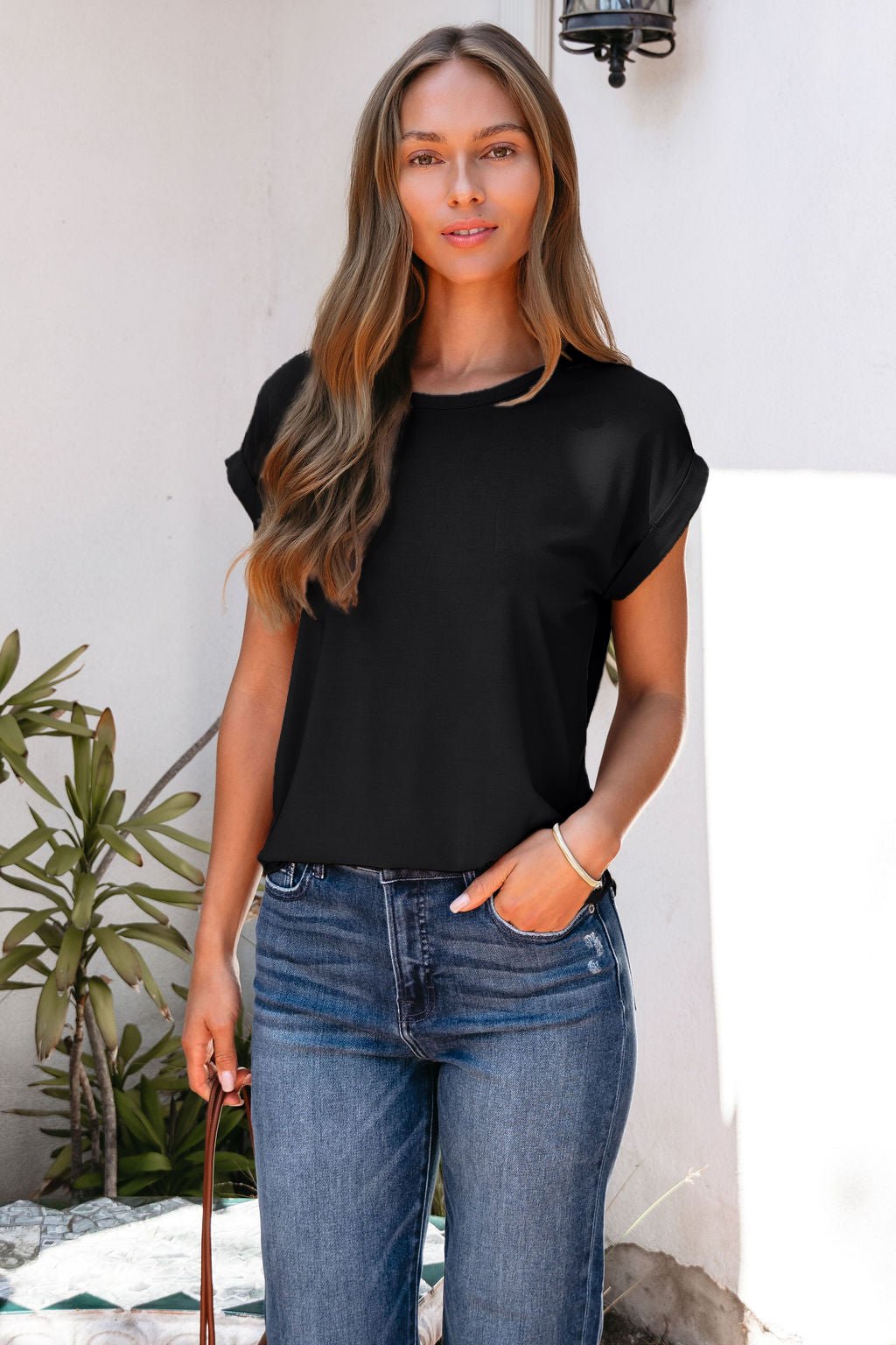 Woman with long light brown hair in a black Soft Modal Basic Tee and blue jeans, standing outdoors near a white wall and green plants.