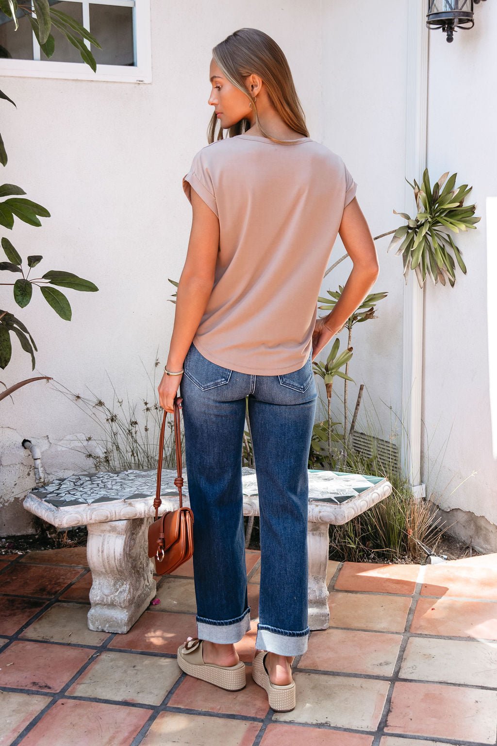 A woman in a relaxed fit Soft Modal Basic Tee - Mocha, blue cuffed jeans, and sandals stands outside with a brown purse, back to camera.