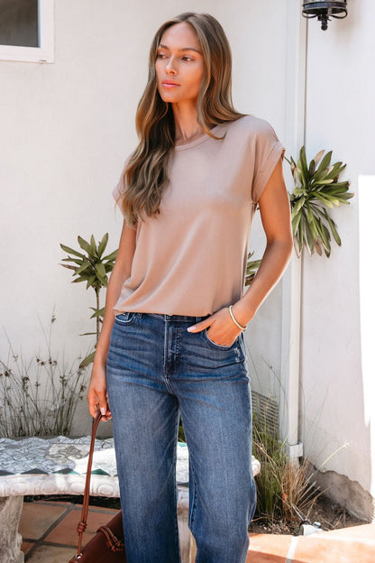 A woman with long hair in a relaxed-fit Soft Modal Basic Tee - Mocha and blue jeans stands by plants, holding a brown bag.