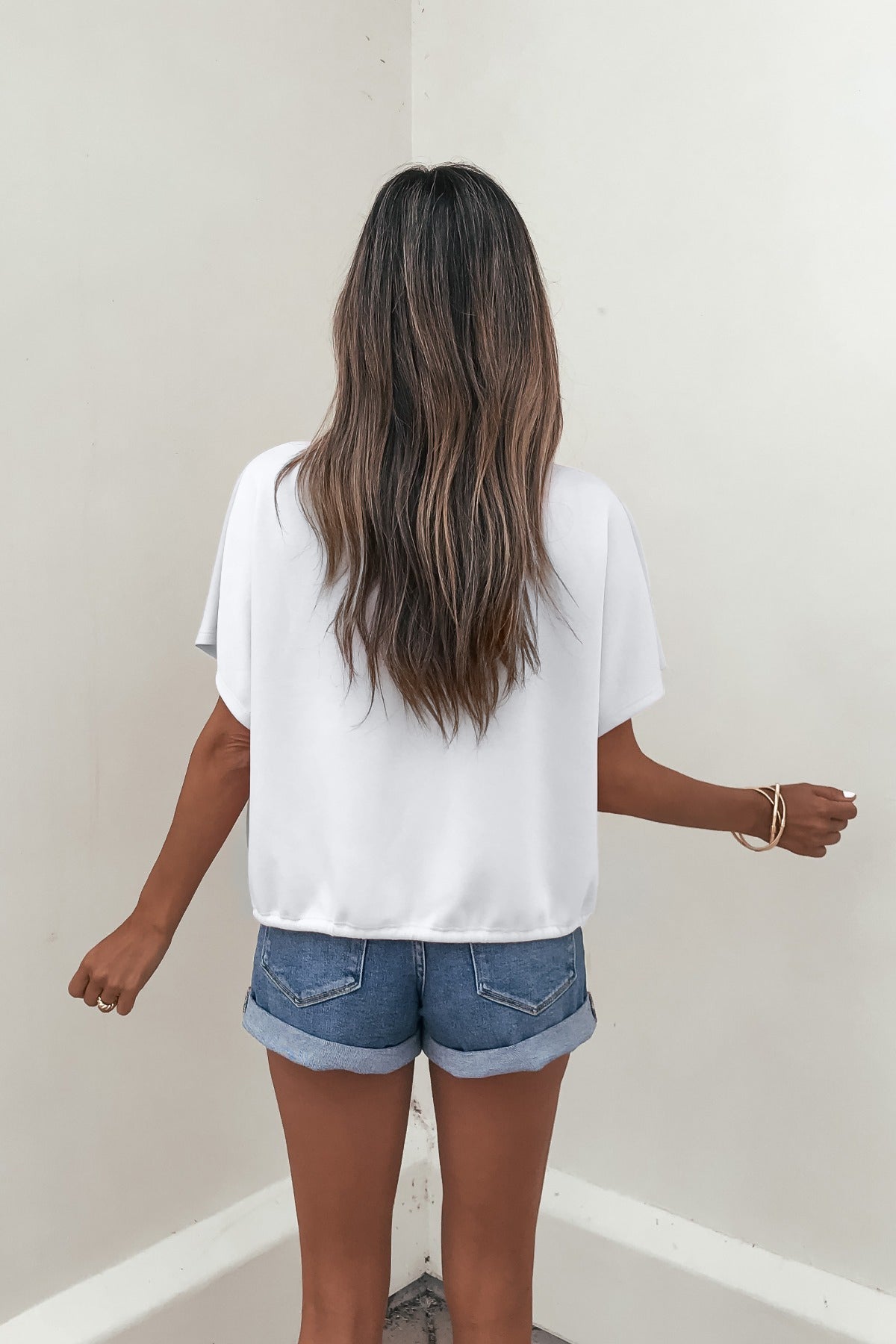 A woman in the Soft Cloud Scuba Lounge Top - White and denim shorts stands facing a light wall, arms relaxed at her sides.