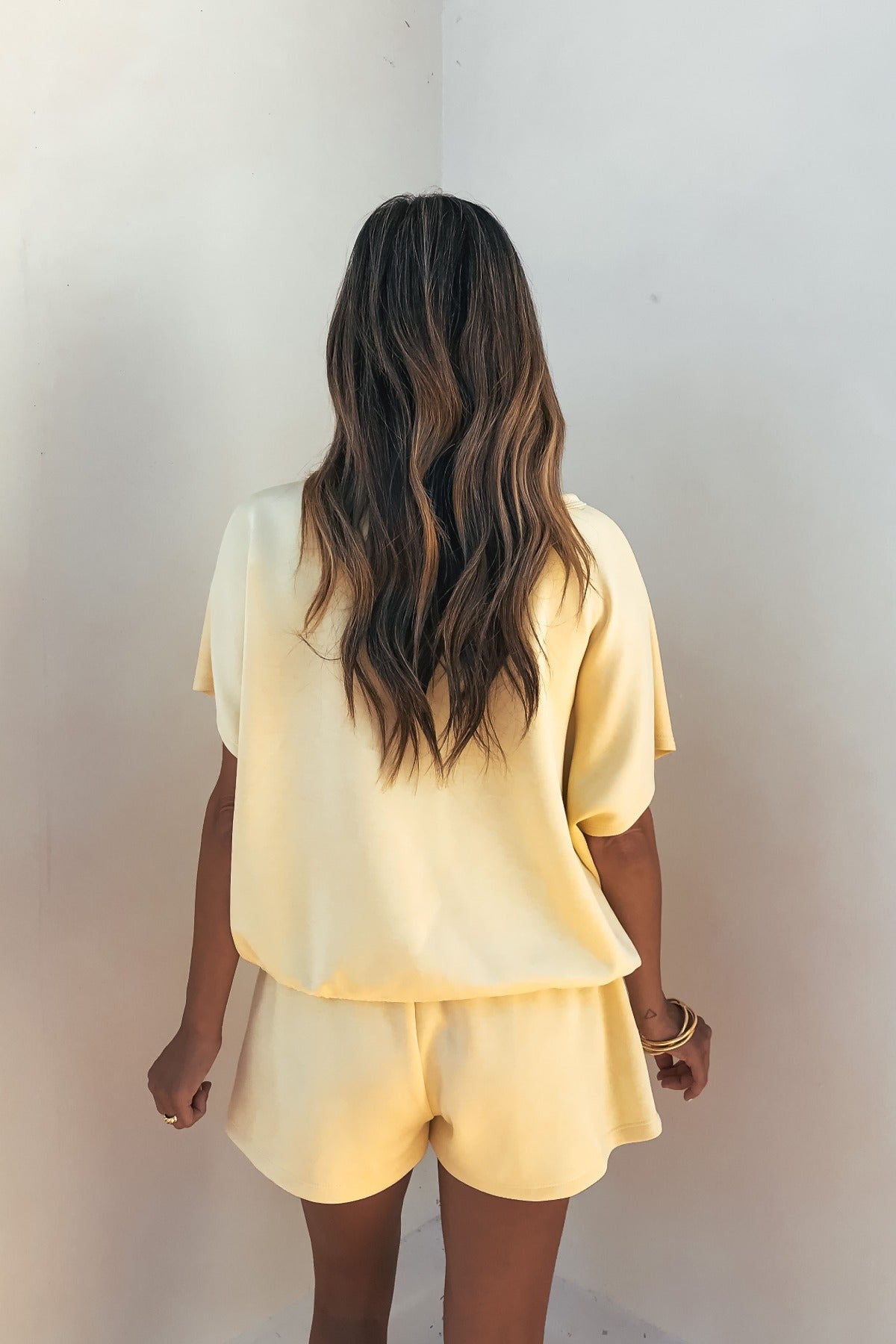A woman with wavy hair faces a white wall, wearing the Soft Cloud Scuba Lounge Top in yellow and matching shorts.