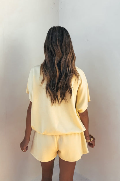 A woman with wavy hair faces a white wall, wearing the Soft Cloud Scuba Lounge Top in yellow and matching shorts.