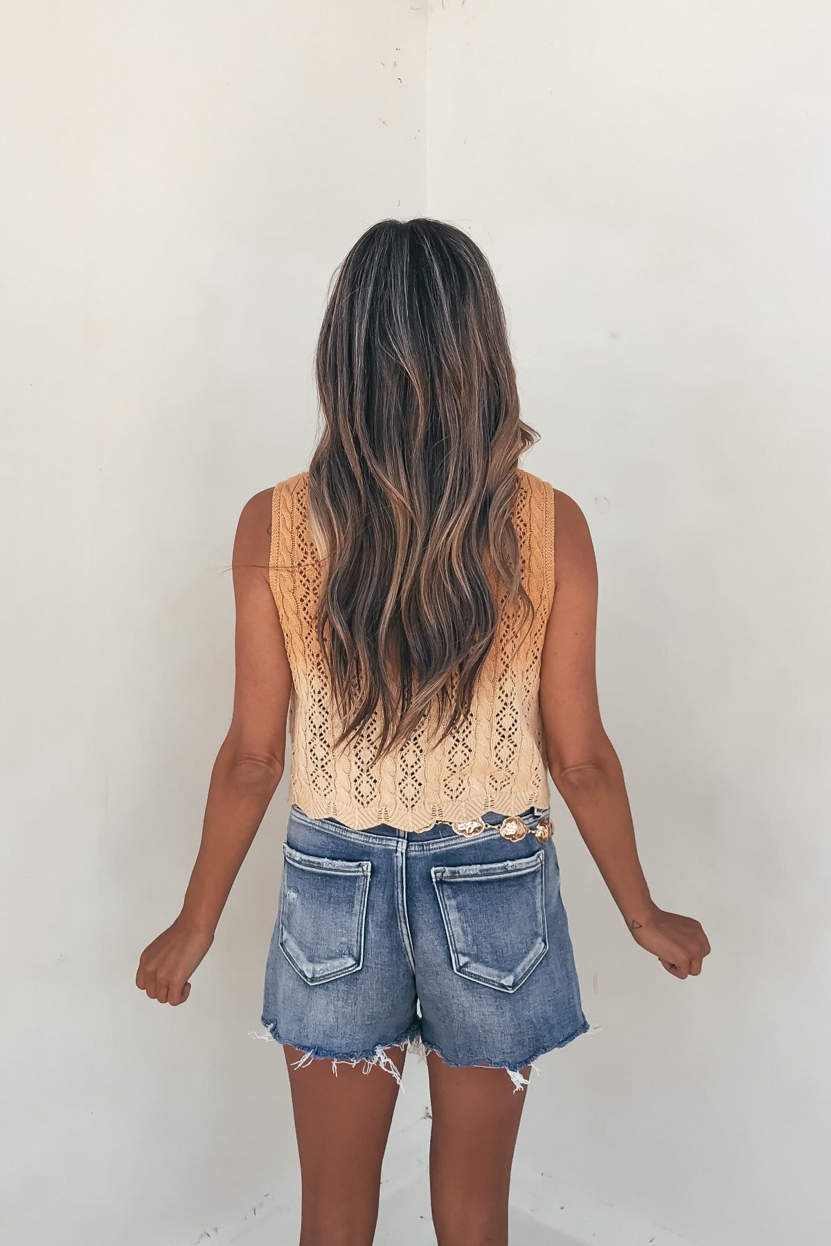 Woman with long wavy hair faces away in the Solstice Ombre Crochet Tie‑Front Top and frayed denim shorts—effortless beach style.