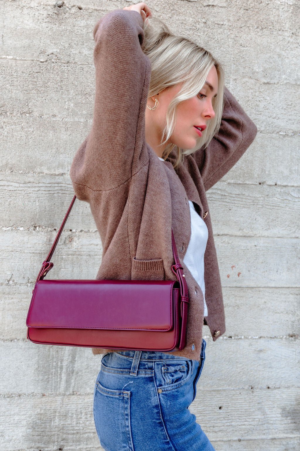 A woman in a brown sweater and jeans poses sideways with the Sorella Burgundy Vegan Leather Shoulder Bag against a concrete wall.