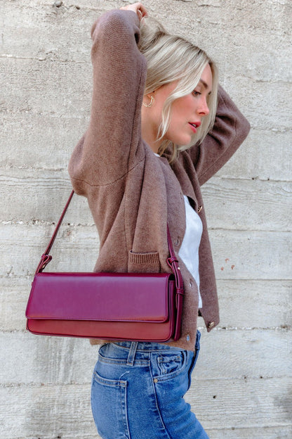 A woman in a brown sweater and jeans poses sideways with the Sorella Burgundy Vegan Leather Shoulder Bag against a concrete wall.