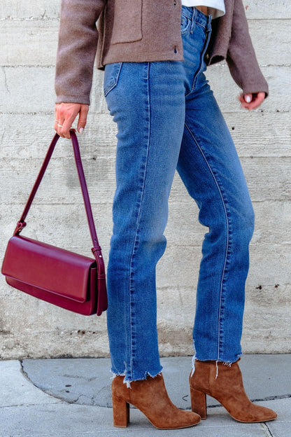 Person in blue jeans, brown boots, a brown jacket, and carrying the Sorella Burgundy Vegan Leather Shoulder Bag against a concrete wall.