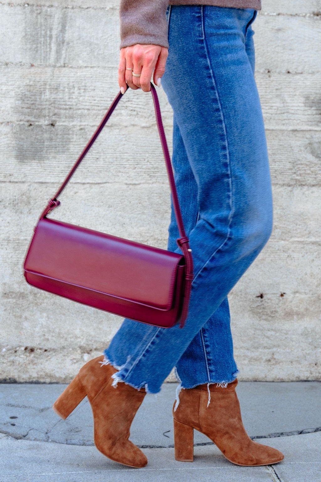 Person carrying the Sorella Burgundy Vegan Leather Shoulder Bag walks past a concrete wall in blue jeans and brown heeled boots.