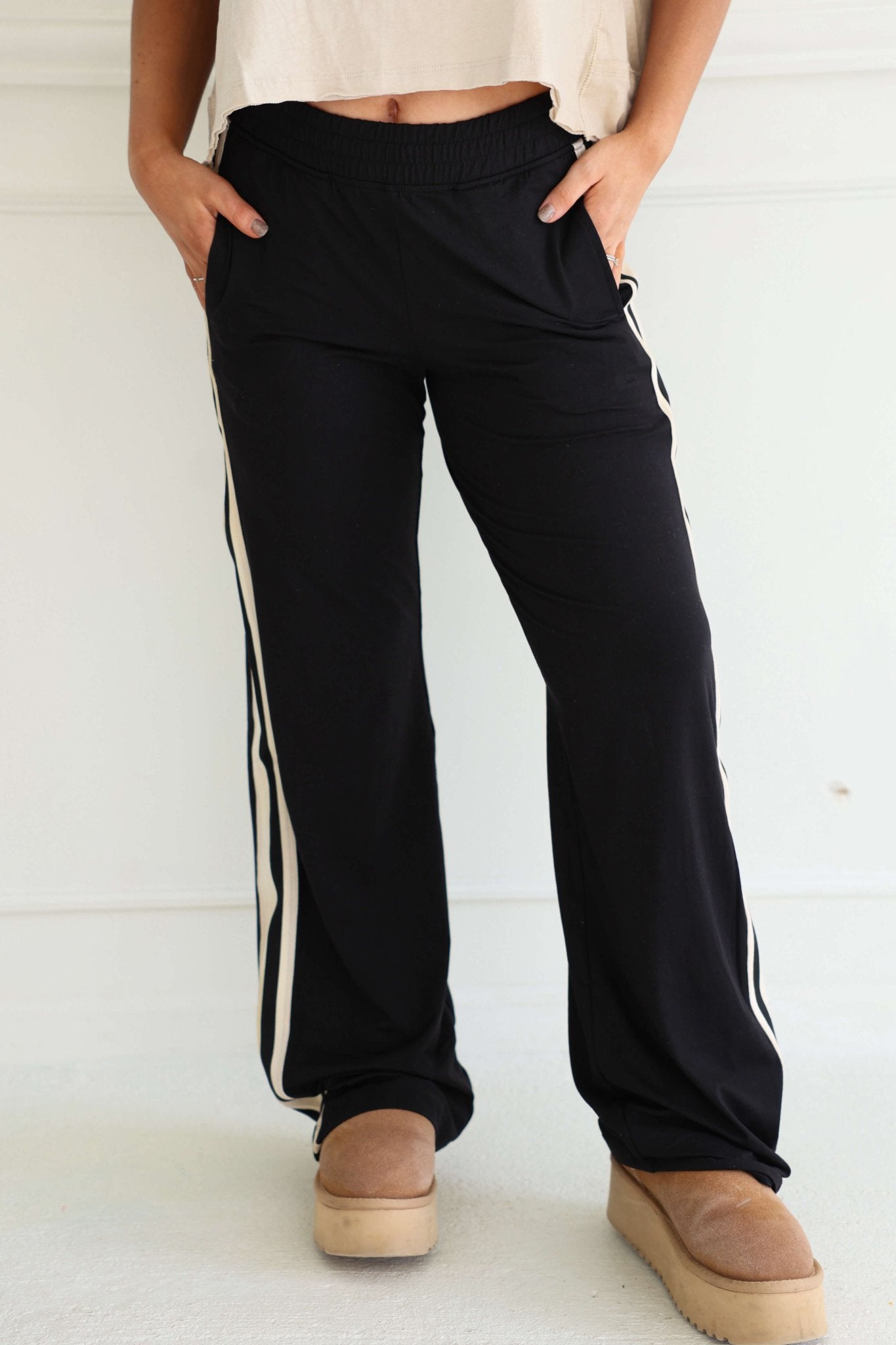 Wearing Sporty Black Side Stripe Pants, a white top, and tan platform slippers, a person stands with hands in pockets.