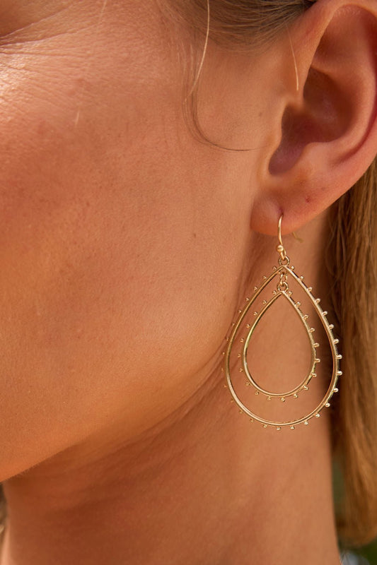 Close-up of a woman's ear wearing St Tropez Dangle Earrings - Gold, with large teardrop shapes and small decorative beads.
