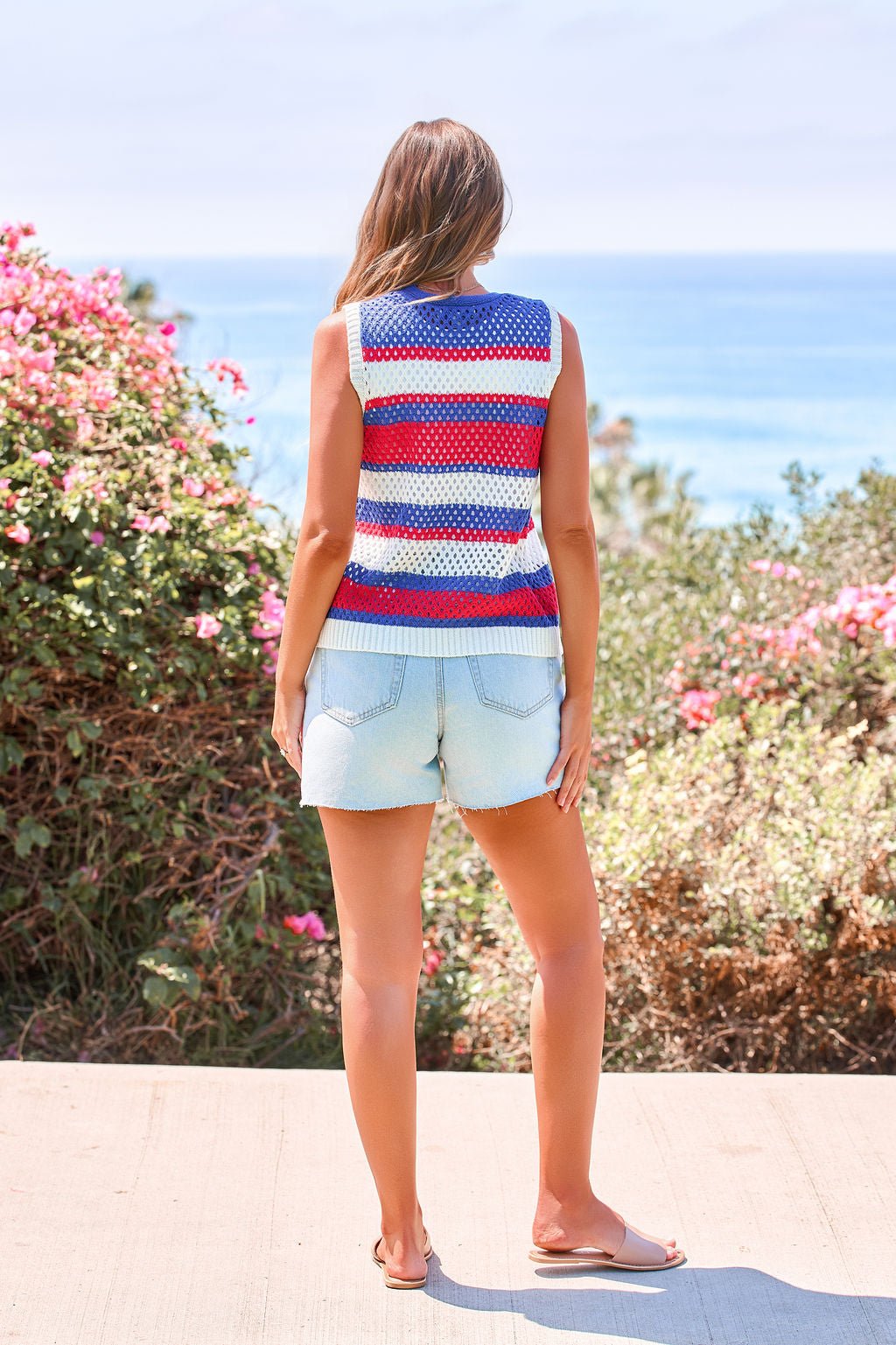 A woman stands by the ocean wearing the Stars & Stripes Tie Front Sweater Vest with light denim shorts and sandals.